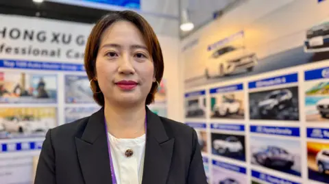Joyce Liu at an EV stall. Joyce has short brown hair and is wearing a white shirt and smart black jacket, facing directly towards the camera. 
