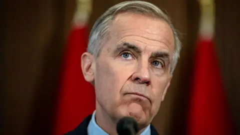 Getty Images A close up image of Mark Carney, who looks out at the distance with a quizzical expression. Behind him is a blurred red background. He is standing at a podium. 