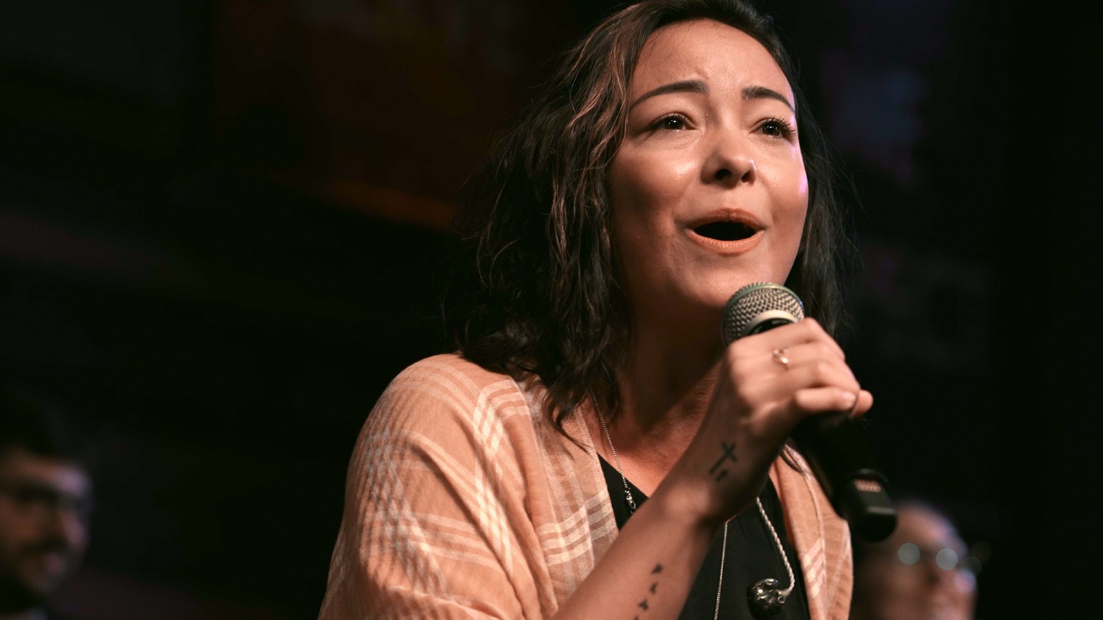 "Woman singing into a microphone on stage with a blurred background, wearing a light shawl, expressive face, indoor concert or performance setting, audience and other musicians faintly visible."