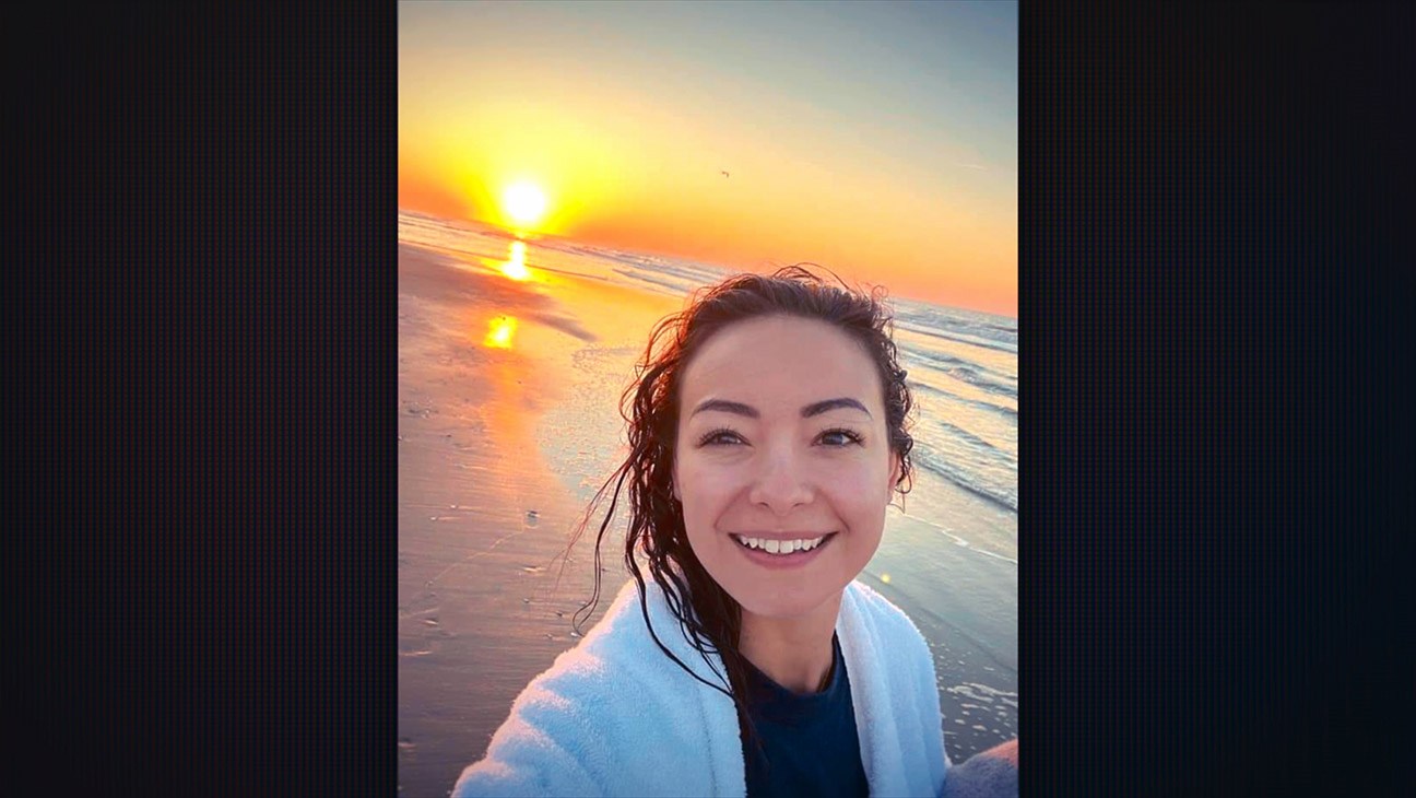 "Smiling woman taking a selfie on a beach at sunrise, wearing a white towel, with wet hair, the sun low over the ocean, and waves gently rolling onto the shore under a colorful sky."
