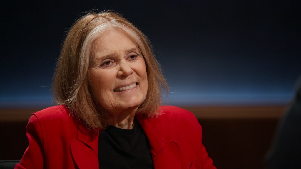 "Smiling elderly woman with shoulder-length light hair, wearing a red blazer and black shirt, sitting indoors in a dimly lit interview or studio setting with a dark, softly illuminated background."